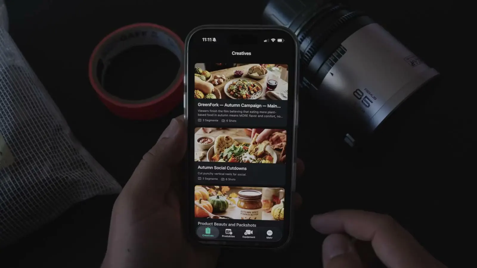 A hand holding an iPhone with the TillyBoard creatives GreenFork Autumn Campaign, Autumn Social Cutdowns and Product Beauty and Packshots. Next to it gaffer tape and an 85 mm cine lens.