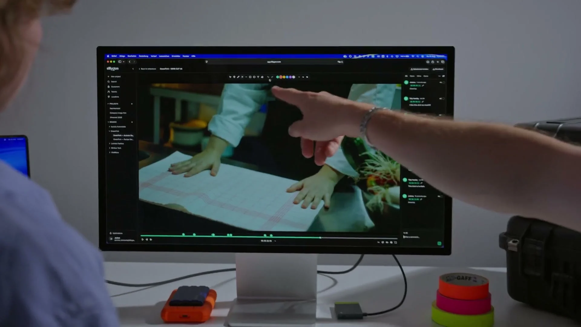 Someone points at a frame in the TillyBoard review player on a large monitor; comment markers sit along the timeline.